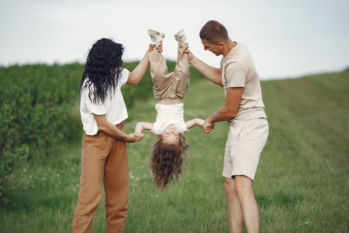 A joyful family enjoys outdoor play, lifting their child upside down in a green field.